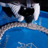 A close-up of a heavily paved silver-toned chain bracelet with a rectangular engraved plate, resting on a bright blue branded velvet surface while a gloved hand holds a black magnifying tool above it.